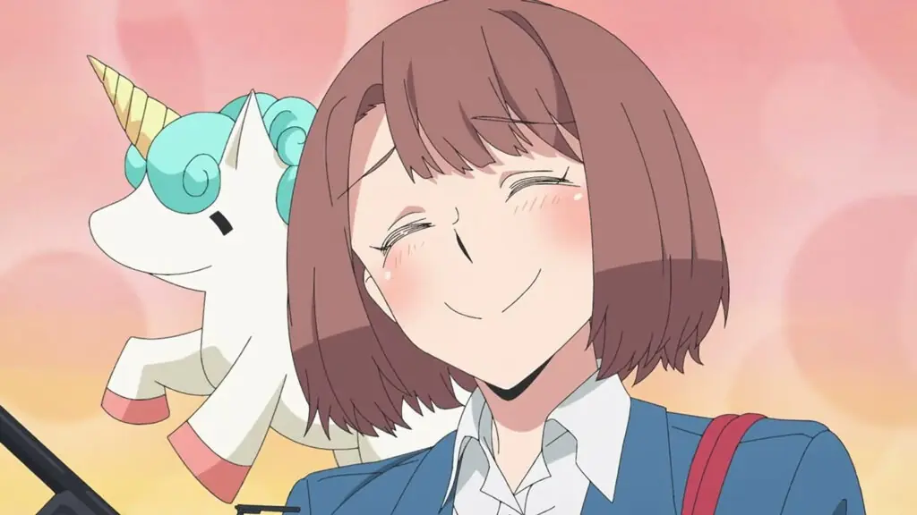 Fotograma del teaser: una joven de pelo corto castaño y chaqueta azul sonríe con los ojos cerrados junto a un peluche de unicornio sobre fondo rosado