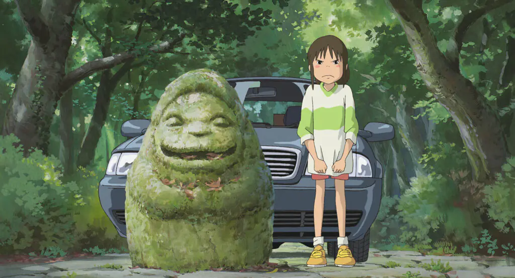 Fotograma de El viaje de Chihiro: una niña con polera blanca y verde, de pie frente a un auto gris, junto a una escultura de piedra cubierta de musgo en un bosque